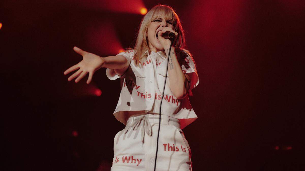 A close-up shot of a woman singing into a microphone. She has blond hair and is dressed in a white short-sleeved top with white pants, both of which have the words quote This is Why repeatedly written in red on them.