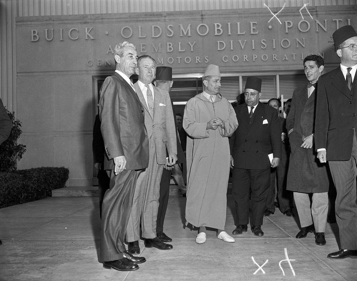 The king, wearing a robe and traditional fez, stands with a group of businessmen in suits in front of the factory. 