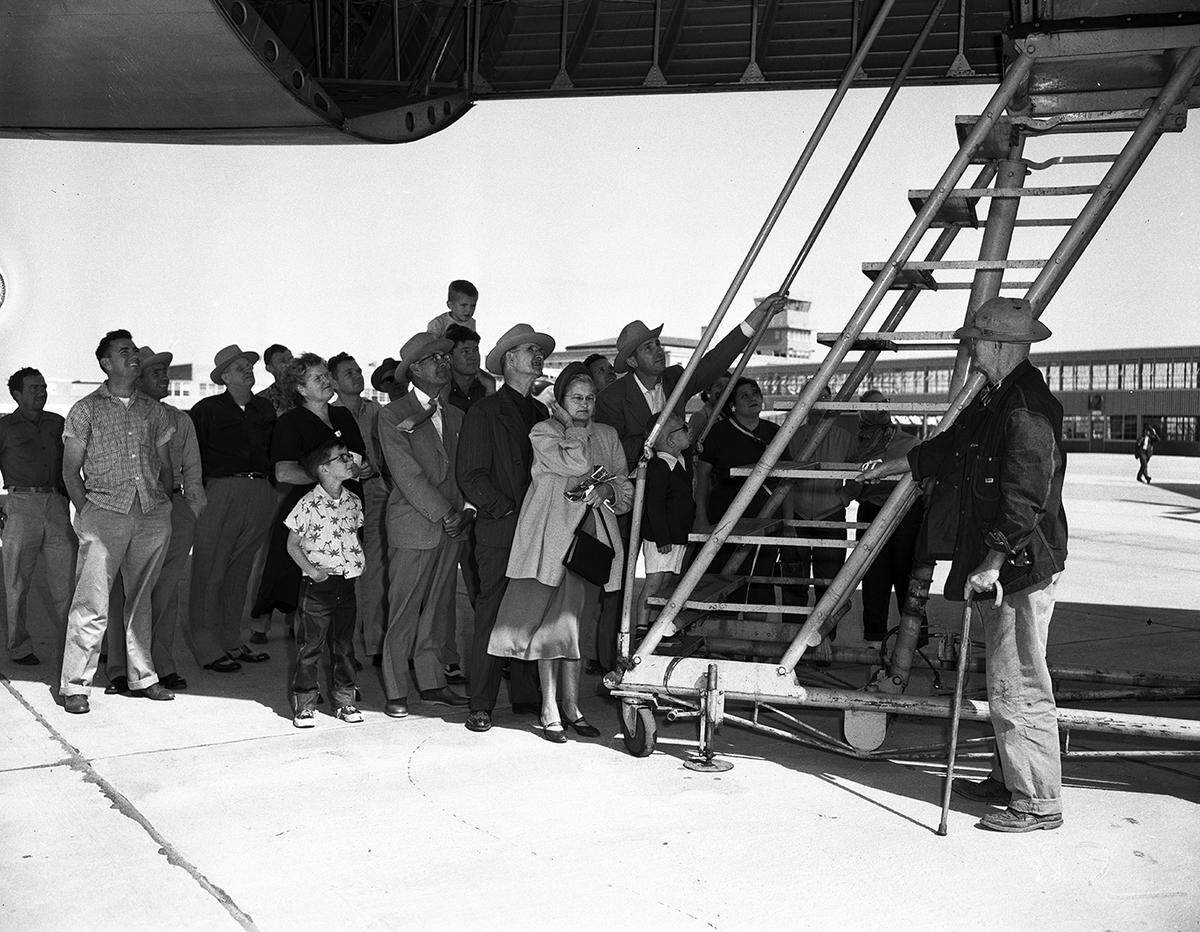 A group of spectators are standing below the belly of an air plane. There is a ladder going up to the opening. The terminal is seen in the background.