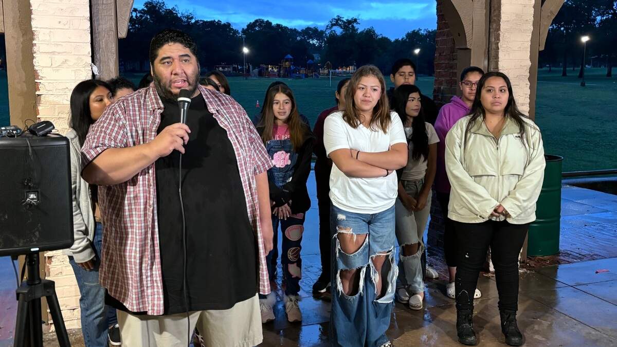 A man in an unbuttoned checkered short sleeve shirt over a black T-shirt stands in front of a group of teenage girls and young women. He speaks into a microphone as dawn breaks in the sky behind them. 