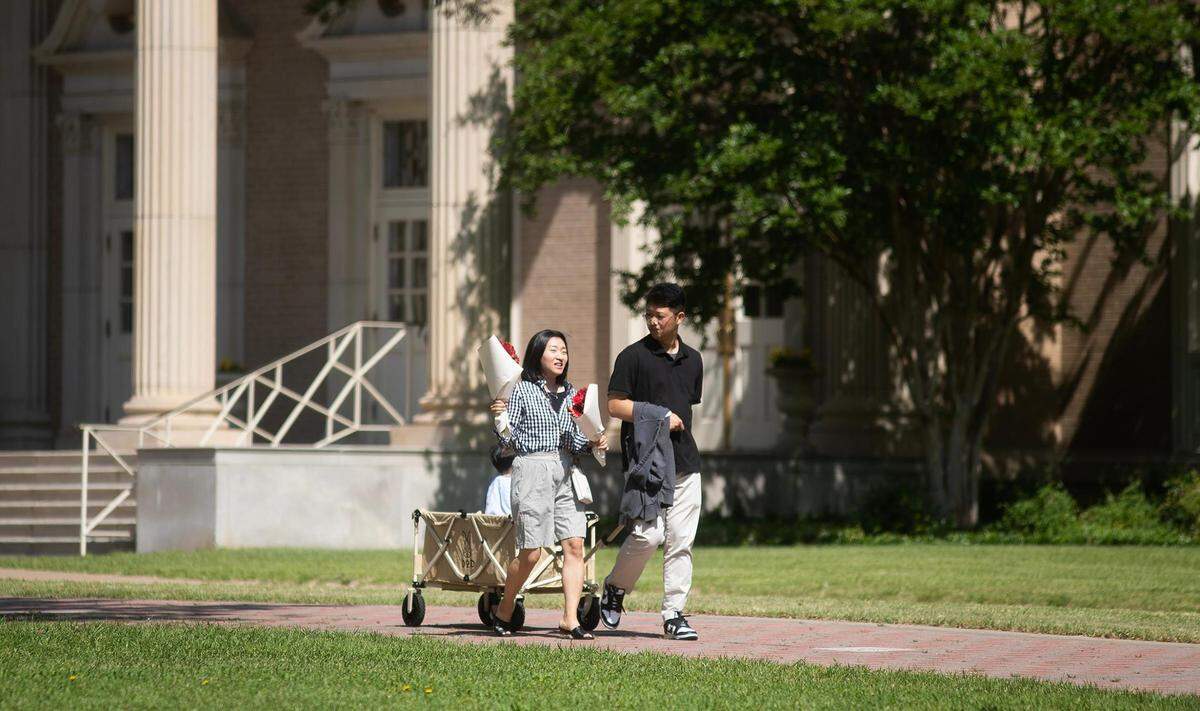 Two people, one holding flowers and the other pulling a wagon, walk down a sidewalk with a campus building behind them.