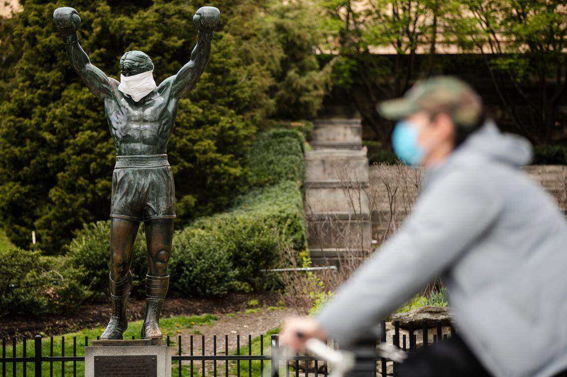A cyclist wearing a protective face mask as a precaution against the coronavirus moves past the Rocky statue outfitted with mock surgical face mask at the Philadelphia Art Museum on Tuesday, April 14, 2020.