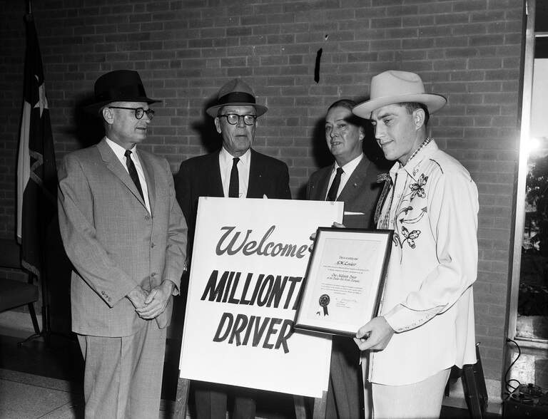E. W. Locker stands at right holding a framed award beside three men and a sign welcoming the millionth driver.