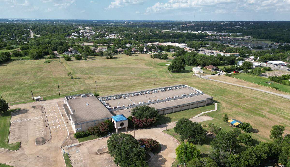 A drone photograph with a one-story concrete building in the foreground covered by open grass. In the distance trees and neighborhoods can be seen.