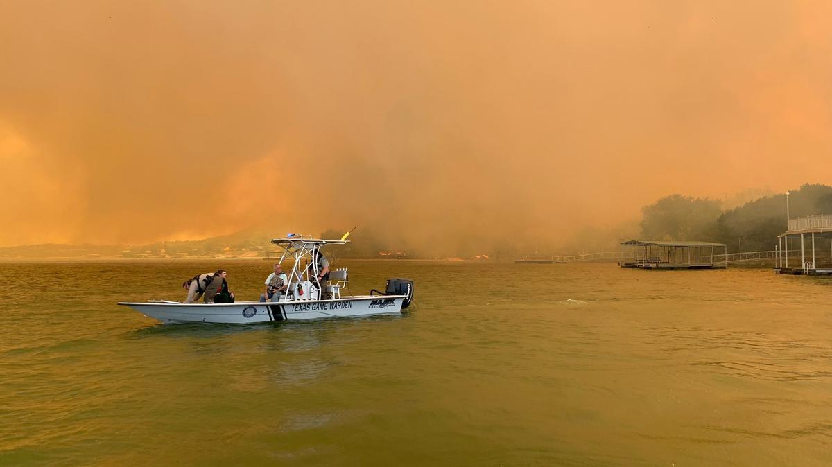 A game warden boat with four people floats on a lake in Palo Pinto County. The smoke filled sky in the background is orange from the wildfires.