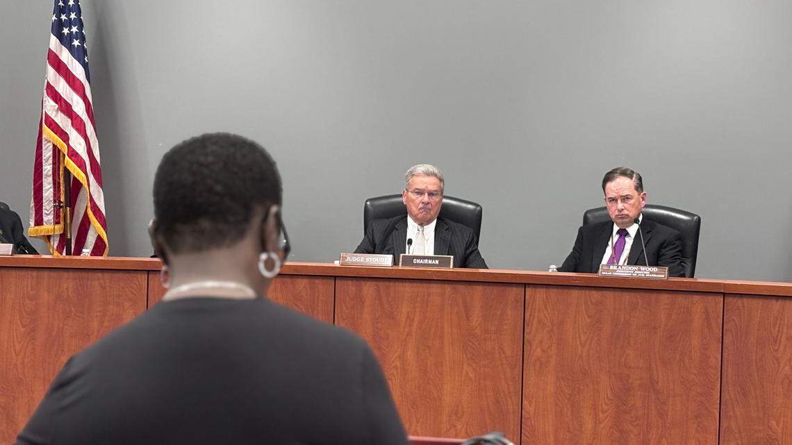Two middle-aged white men in dark suits are in focus seated at a dais in a grey room. A Black woman out of focus in the foreground faces them as they listen to her speak. An American flag stands at the left of the frame behind the men.