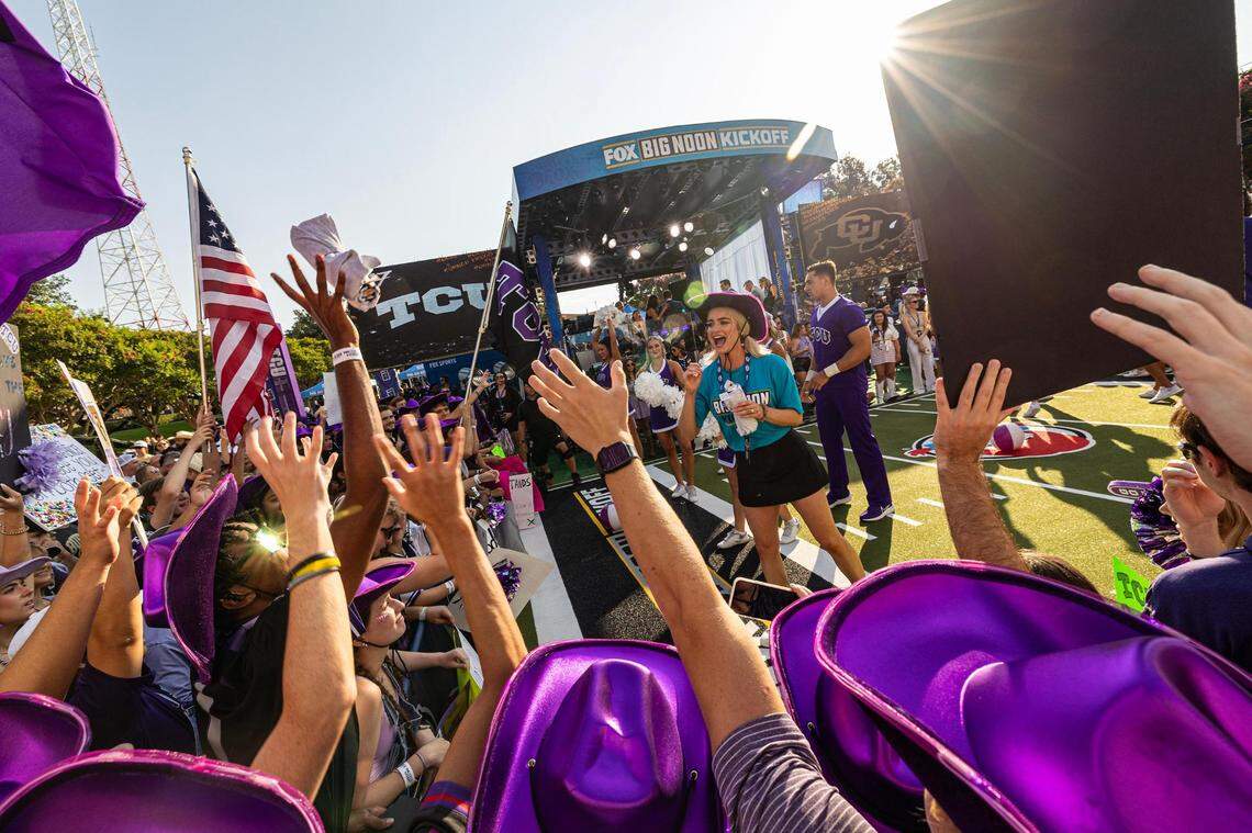 Hands reach into the air, many people wearing purple cowboys hats, to catch a t-shirt being thrown while the Fox Big Noon Kickoff stage is in the background.