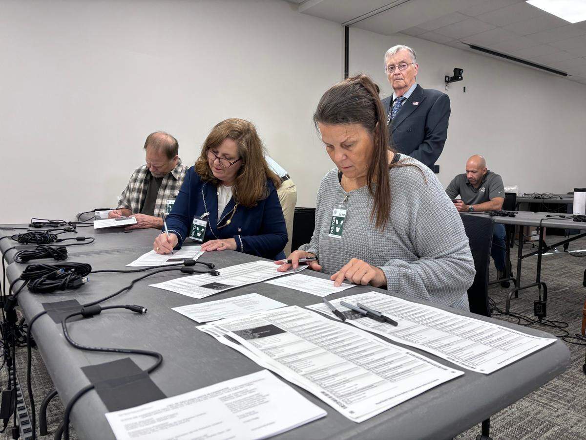 A woman in a grey sweater fills out a form on a table. A woman in a blue jacket does the same seated next to her, while a man with white hair and glasses stands behind them watching.