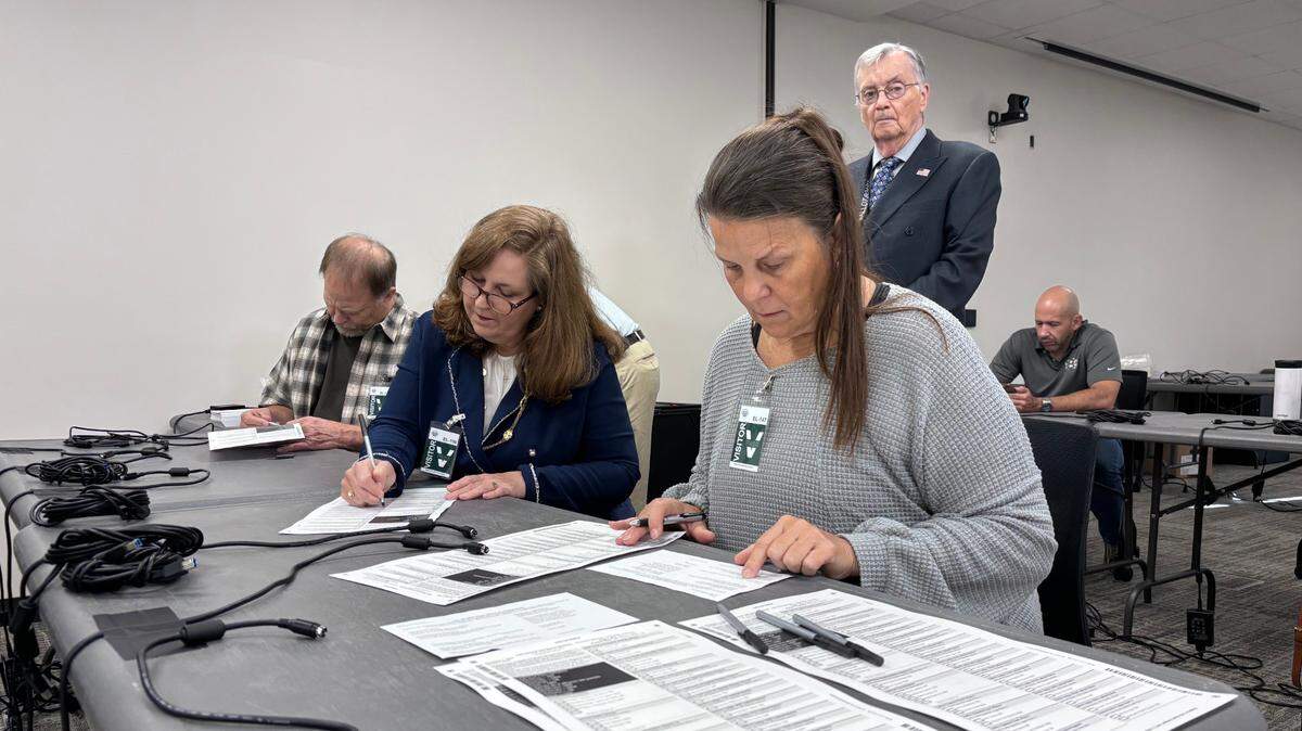 A woman in a grey sweater fills out a form on a table. A woman in a blue jacket does the same seated next to her, while a man with white hair and glasses stands behind them watching.