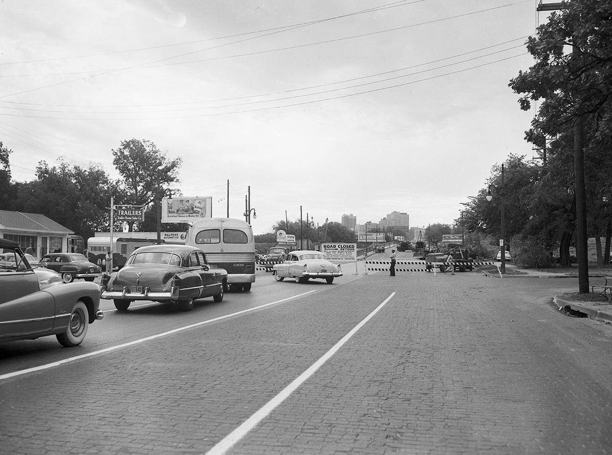 Black and white photo taken in 1953. About four automobiles are seen in line in front of a barrier blocking their way to cross the West 7th Street bridge in Fort Worth heading into downtown.