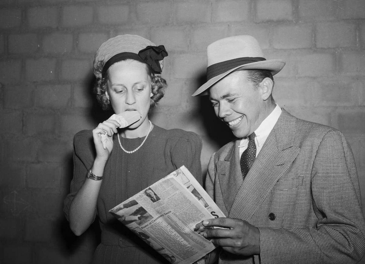 Miss Weir is wearing a nice dress, a necklace, a hat, and a watch while Mr. Greenwood is wearing a suit, tie, and hat. Miss Weir is eating an ice cream sucker and looking at the player lineup that Mr. Greenwood is holding in his hands. They are standing in front of a brick structure.