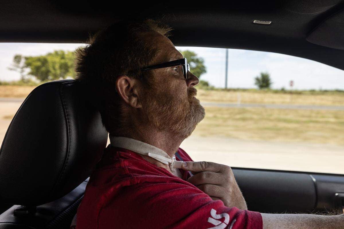 Head and shoulder profile view of Kevin driving his car on a rural road. He is touching his tracheostomy with his left hand.