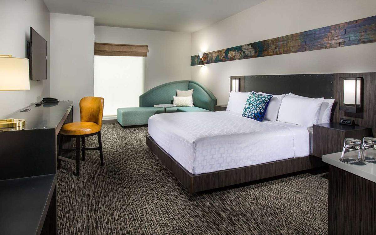 A Cambria hotel room shows a queen size bed, sofa, desk and chair, TV and mini bar. 