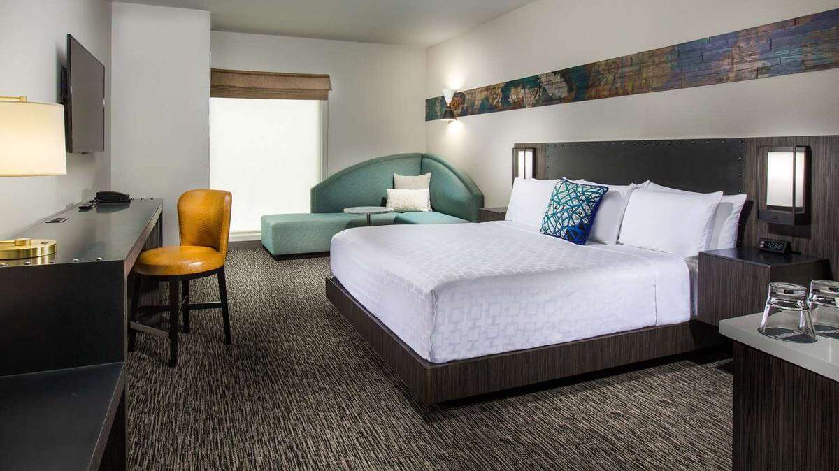 A Cambria hotel room shows a queen size bed, sofa, desk and chair, TV and mini bar. 