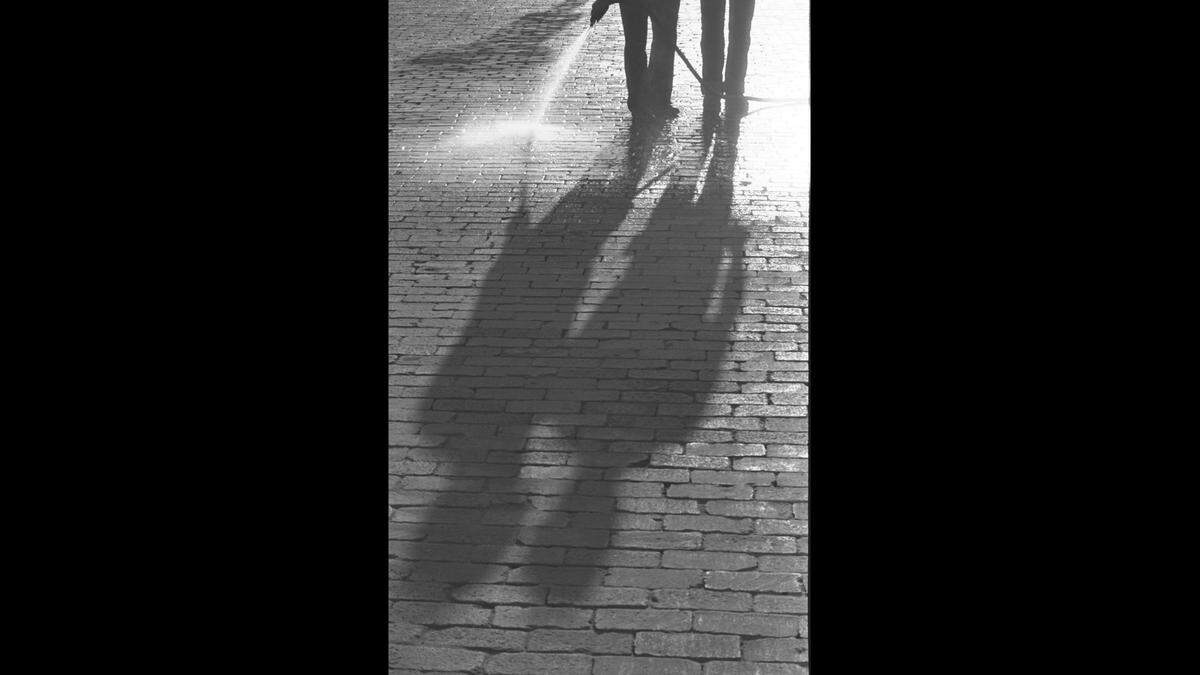 Two men cast shadows over bricks on Camp Bowie Boulevard. One man to the left is holding a hose and spraying water on the bricks.