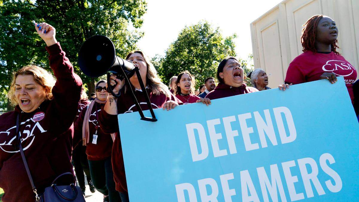 A crowd of protesters hold up a large sign that says 'Defend Dreamers.' One protester is shouting into a megaphone.