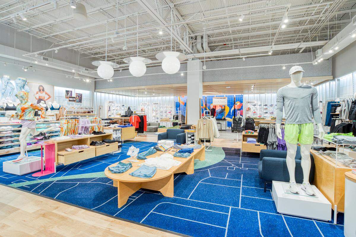 A Nike Live floor features men's clothing on the right and women's clothing on the left. A male mannequin models a gray shirt and neon green running shorts next to a table of running shorts and hoodies. To the left, a female mannequin models purple shorts and a tie-dyed hoodie.  