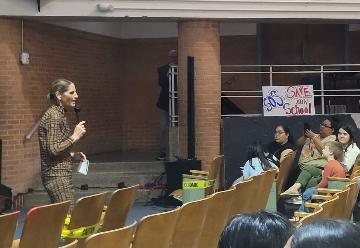 A woman stands in front of a crowd and speaks. To the side, a sign reading "SOS Save Our Schools" hangs from a railing.