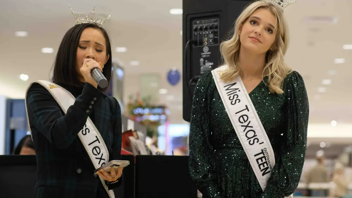 Two women in black, sleeved dresses stand next to each other in a Texas shopping mall. The woman on the left is singing into a micophone and wearing a sash that says quote Miss Texas. The woman on the right is looking at the audience, wearing a sash that says quote Miss Texas Outstanding Teen. Both are wearing tiaras.
