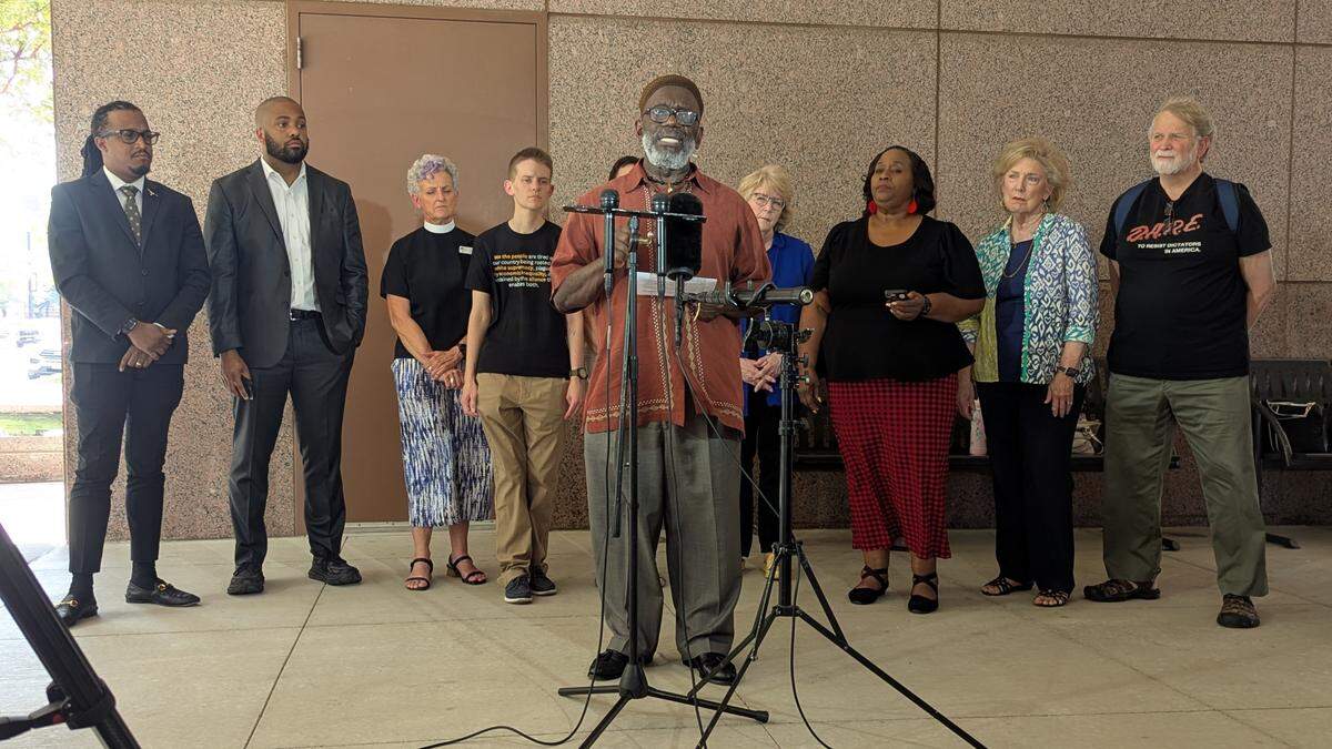 A man stands in front of a podium surrounded by other people while news cameras look on.