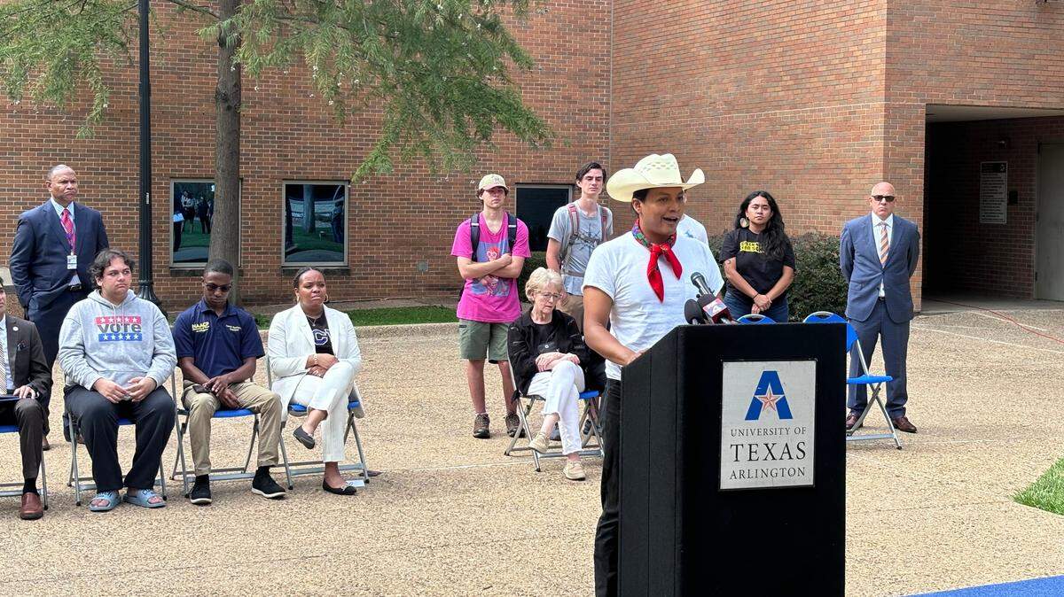 A young man in a white cowboy hat speaks at a lectern set up outside a red brick building. Several people sit in a row of blue chairs behind him.