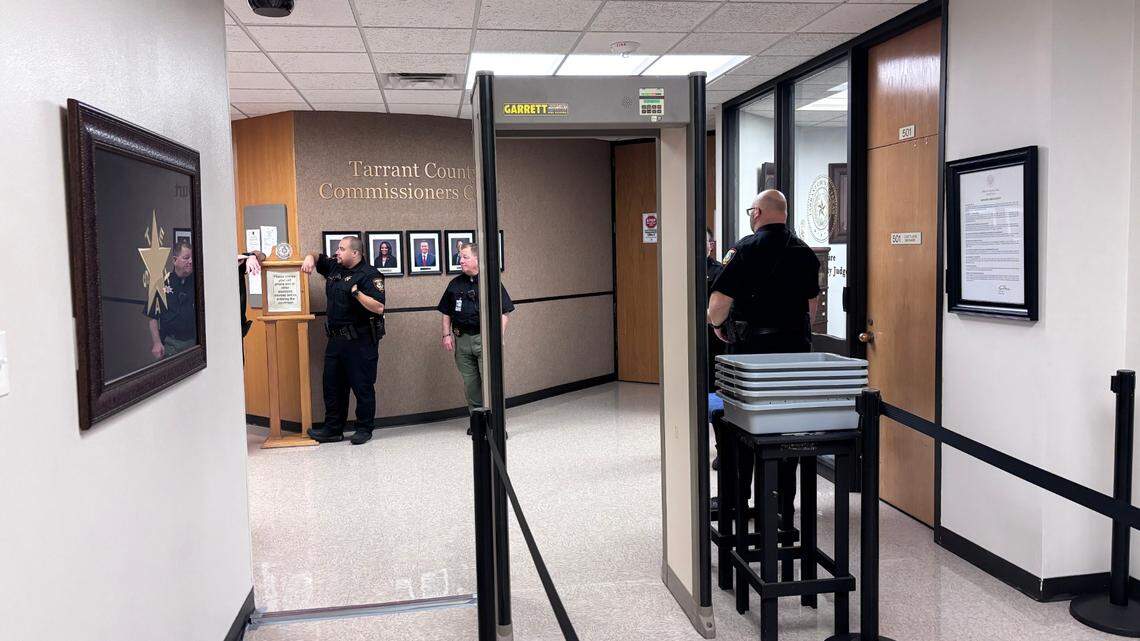 Three law enforcement officers stand near a metal detector in a hall with white floors and walls. Black stanchion crowd control barriers are set up to lead to the detector. 