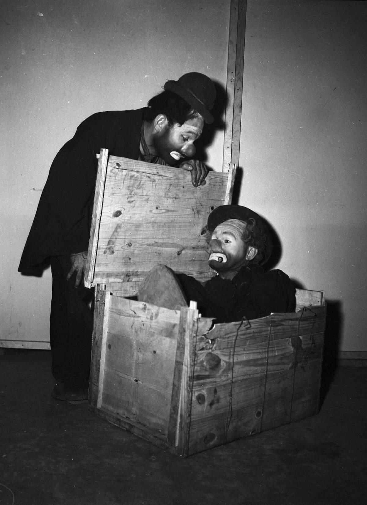 Two men dressed like clowns stare at each other as one opens a large crate where the other is squatting.