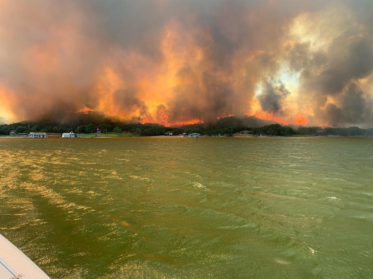 A lake in Palo Pinto County with hills on fire in the background, smoke is rising in the air.