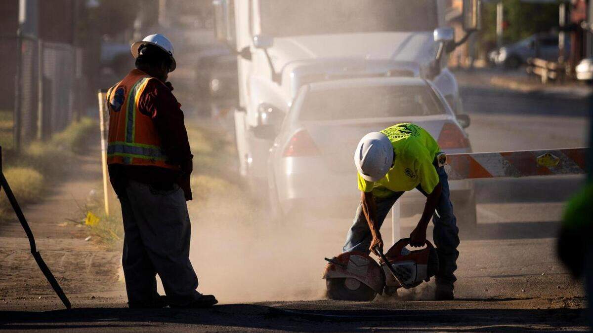 Two construction workers, one wearing a bright orange safety vest and one wearing a neon green shirt, work outside, with the skyline of Fort Worth visible behind them and a cloud of dust coming from their work.