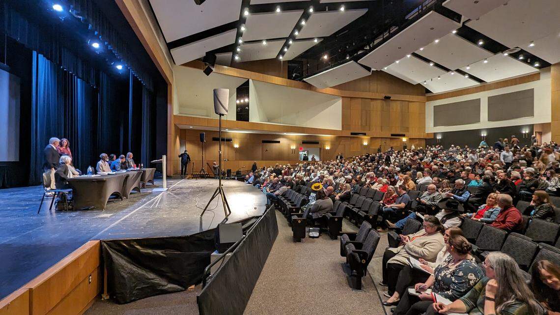 A wide shot of an auditorium with a panel of speakers to the left and an audience of around a thousand to the right. 