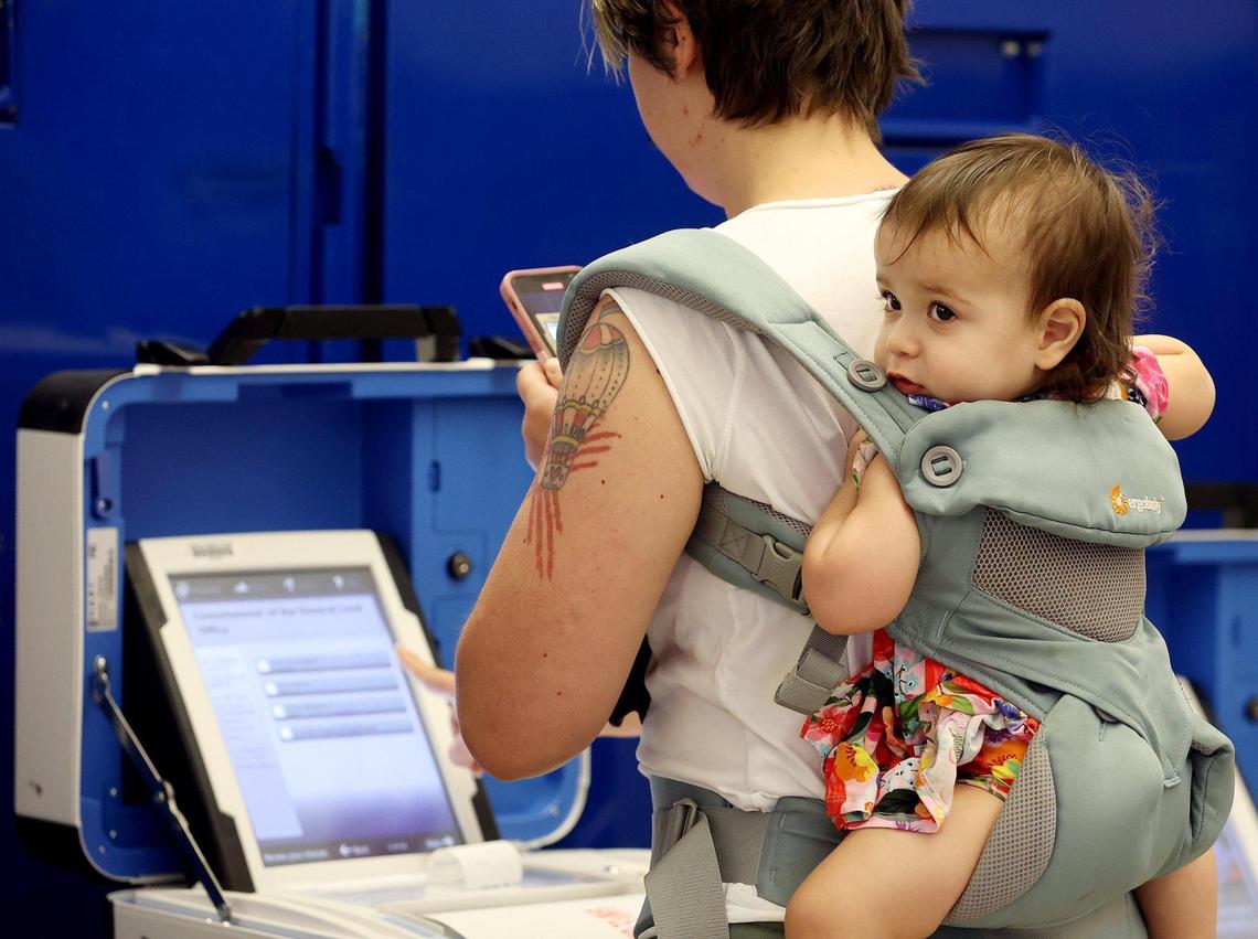 Tristan Martinez takes a video of how to fill out an in-person ballot on a voting machine while her daughter, Eliana, 1, waits patiently on her back on Friday. The public was invited to test the system and had the opportunity to run in-person and absentee ballots.