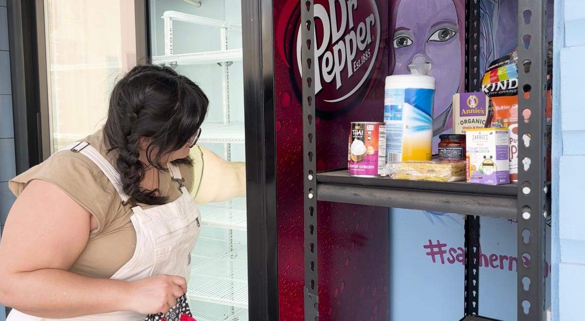 A woman puts food in an outdoor fridge. A stack of food sits on a shelf next to her in front of a painting of Atatiana Jefferson.