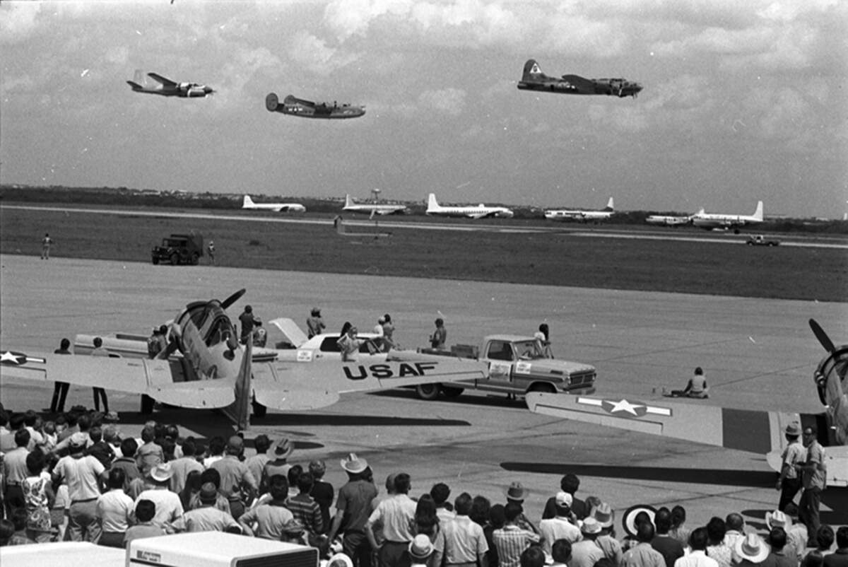 There is a crowd towards the foreground of the photo. There is two planes near the crowd at the front. Three planes seen flying. Six commercial ,white , passenger planes can be seen in the background.
