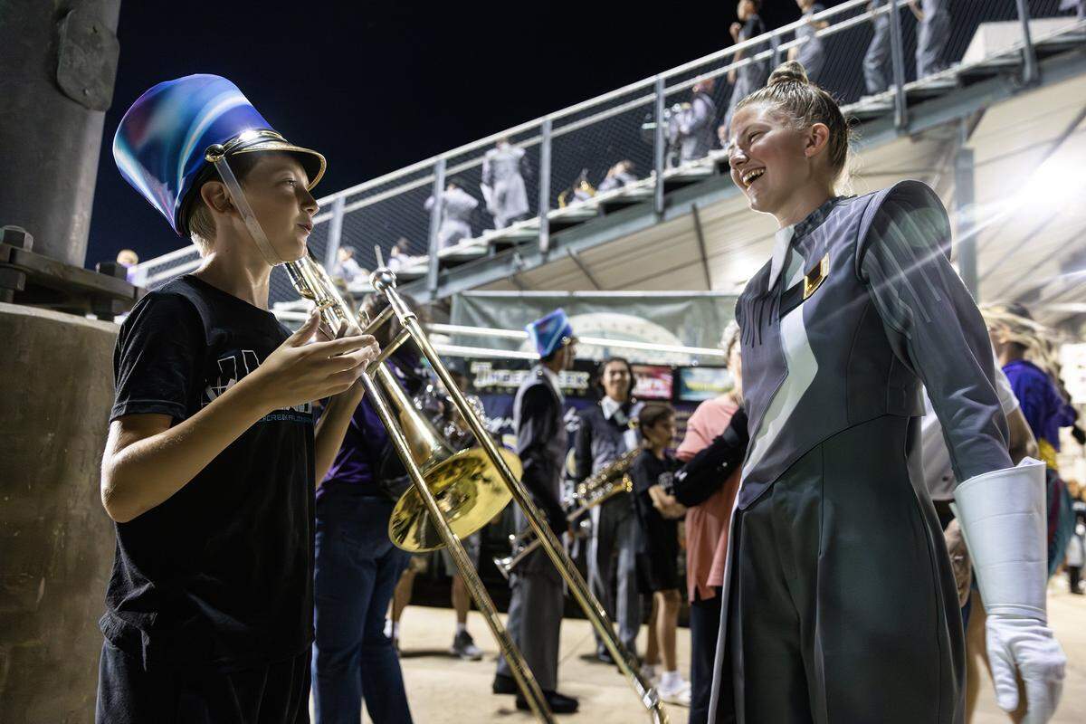 A young boy wearing a marching band hat and holding a trombone smiles at his sister to the left who is wearing a grey band uniform and large white gloves.