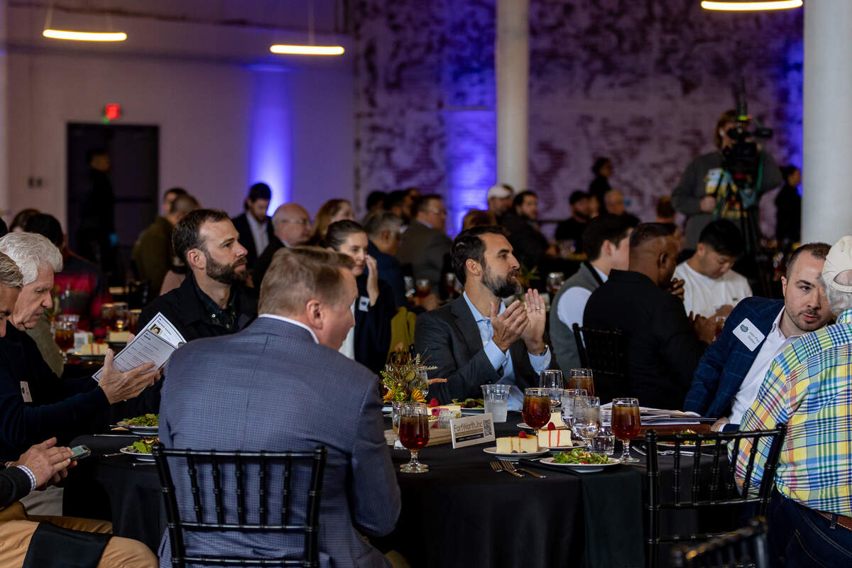 Dozens of people sit around circle tables to eat lunch. The tables have black table clothes, glasses for water and sweet tea and salad plates for lunch. Some guests eat, some look at their event programs and others clap for an event speaker.