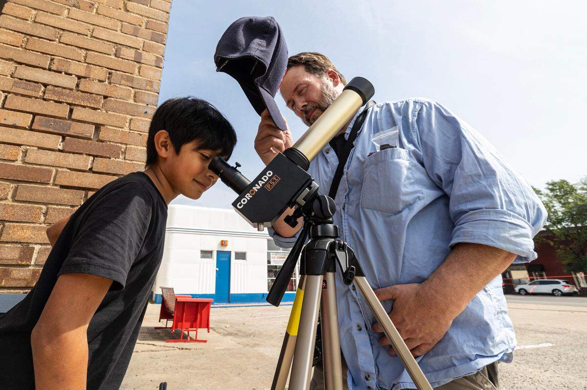 A child looks into a telescope on a tripod on the left while a man holds a baseball cap to block the sun's glare beside him.