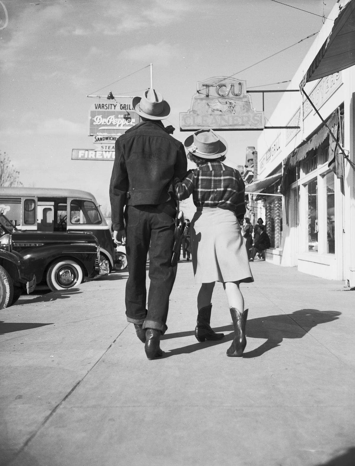 They are photographed from the back, walking on a sidewalk. Mr. Barber is wearing a coat, jeans, cowboy boots and a cowboy hat. His pant legs are cuffed. Miss Reuther is wearing a plaid blouse, a skirt, a cowboy hat and cowboy boots. At the left are several cars parked along the side of the road. To the right are a variety of stores and shops.