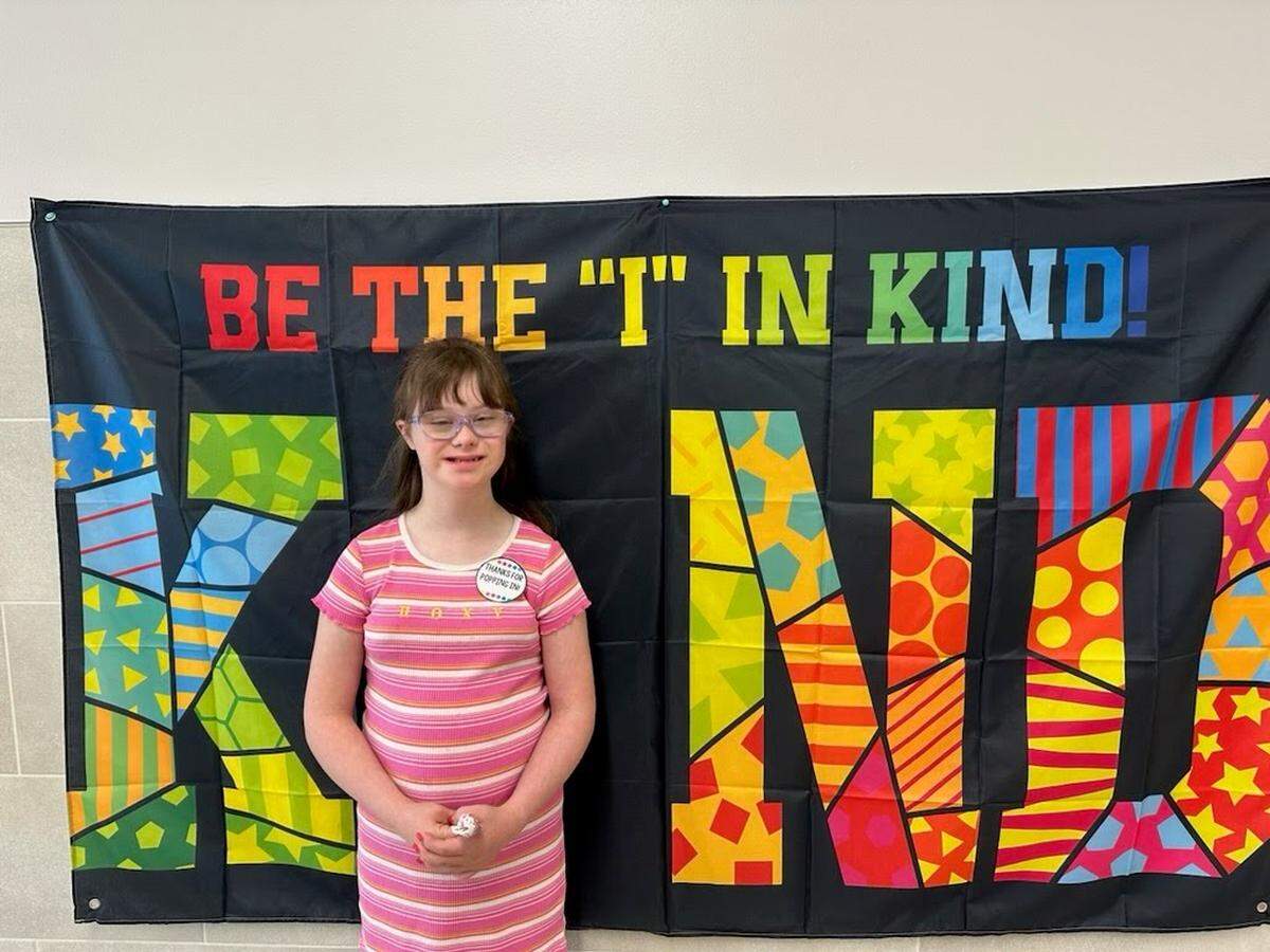 A girl in a pink and white striped dress stands in front of a flag that says “Be the I in Kind.”