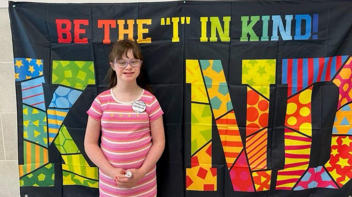 A girl in a pink and white striped dress stands in front of a flag that says “Be the I in Kind.”