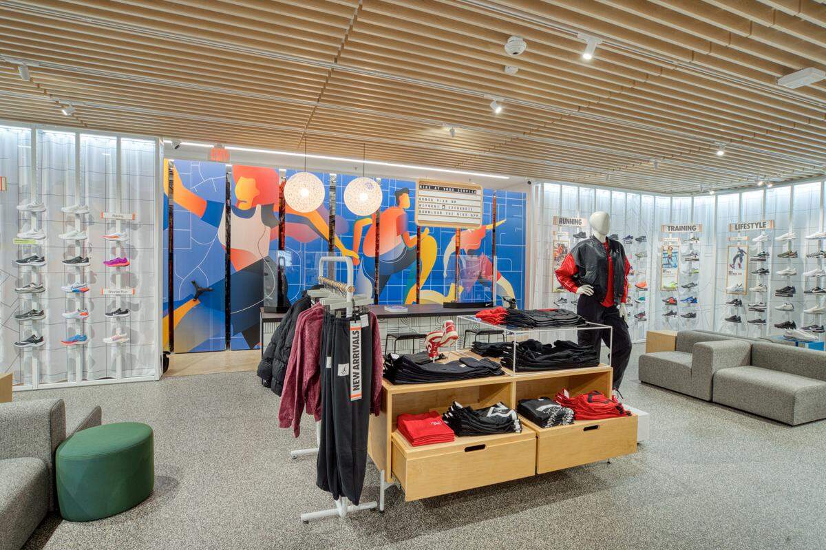 A Nike Live store wall features a large wall with six rows of lifestyle, training and running shoes. Next to the wall sits four gray couches for guests to sit and try on shoes. The area also features a table with thick running jackets and sweatpants. The back wall of the display is decorated with abstract art of people running and exercising.