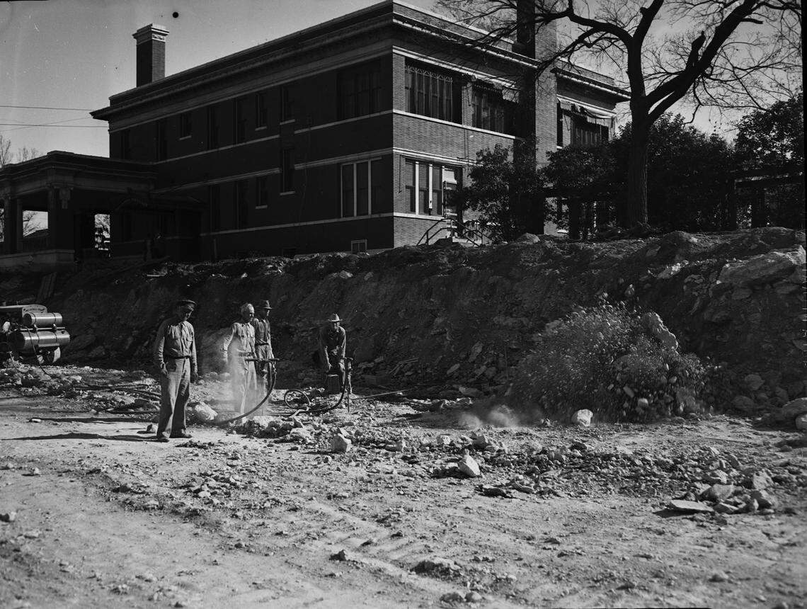 Dec. 3, 1941: Cutting through rock at top of West Lancaster Avenue bluff for the cut and fill project to connect Trinity Bridge with Summit Avenue, three workmen cut loose a blast of eight sticks of dynamite. At right, J. W. Suritt bends over the battery which sets off a charge buried deep in rock. Above the bluff is a brick building with many windows and few trees. Behind the workmen is some construction equipment.