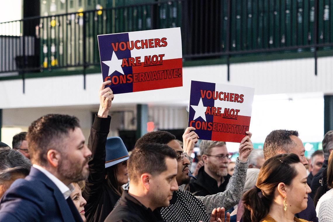 Event attendees hold signs saying “Vouchers are not conservative” while Gov. Greg Abbott speaks during March 6 at Temple Christian School in Fort Worth.