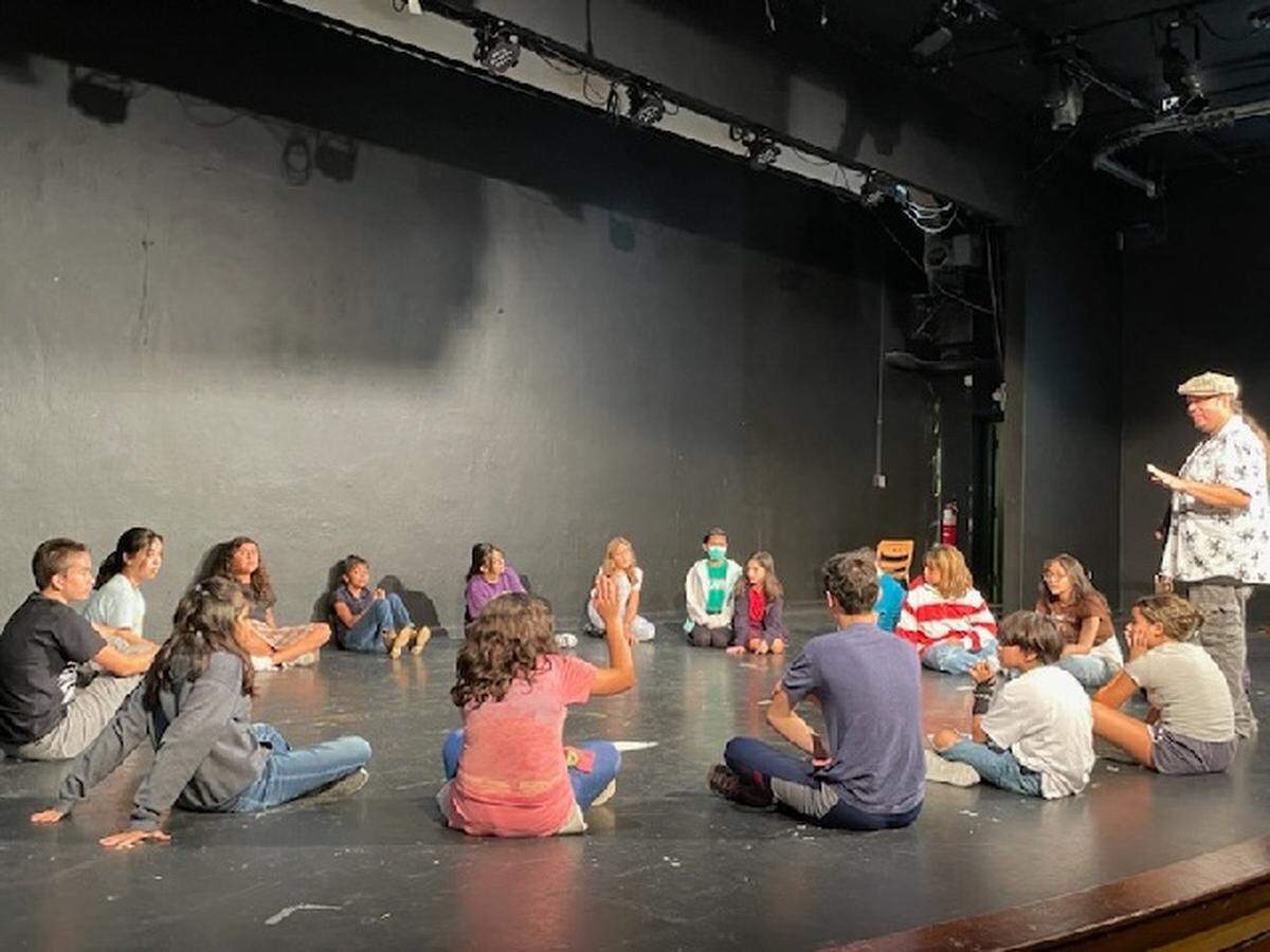 A man stands on stage speaking to a group of youth actors seated in a circle on the stage floor.