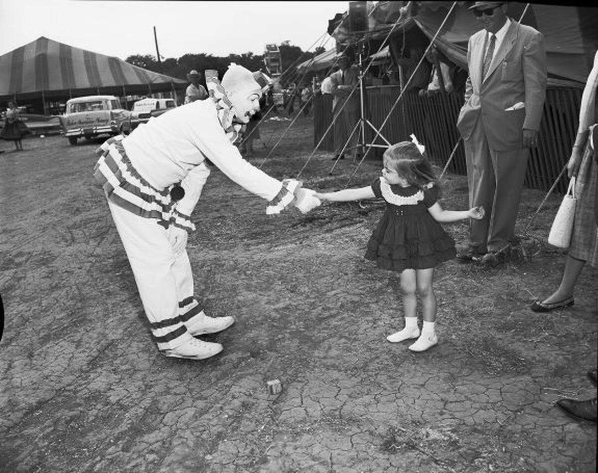 A hesitant looking girl shakes the hand of a clown bending down to say hello.