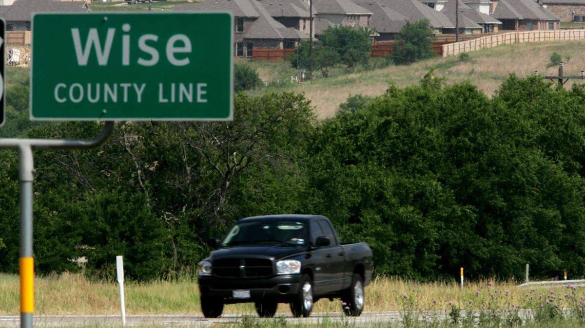 A pickup truck drives down a rural road with new houses seen in the background. A Wise County line sign is in the foreground.