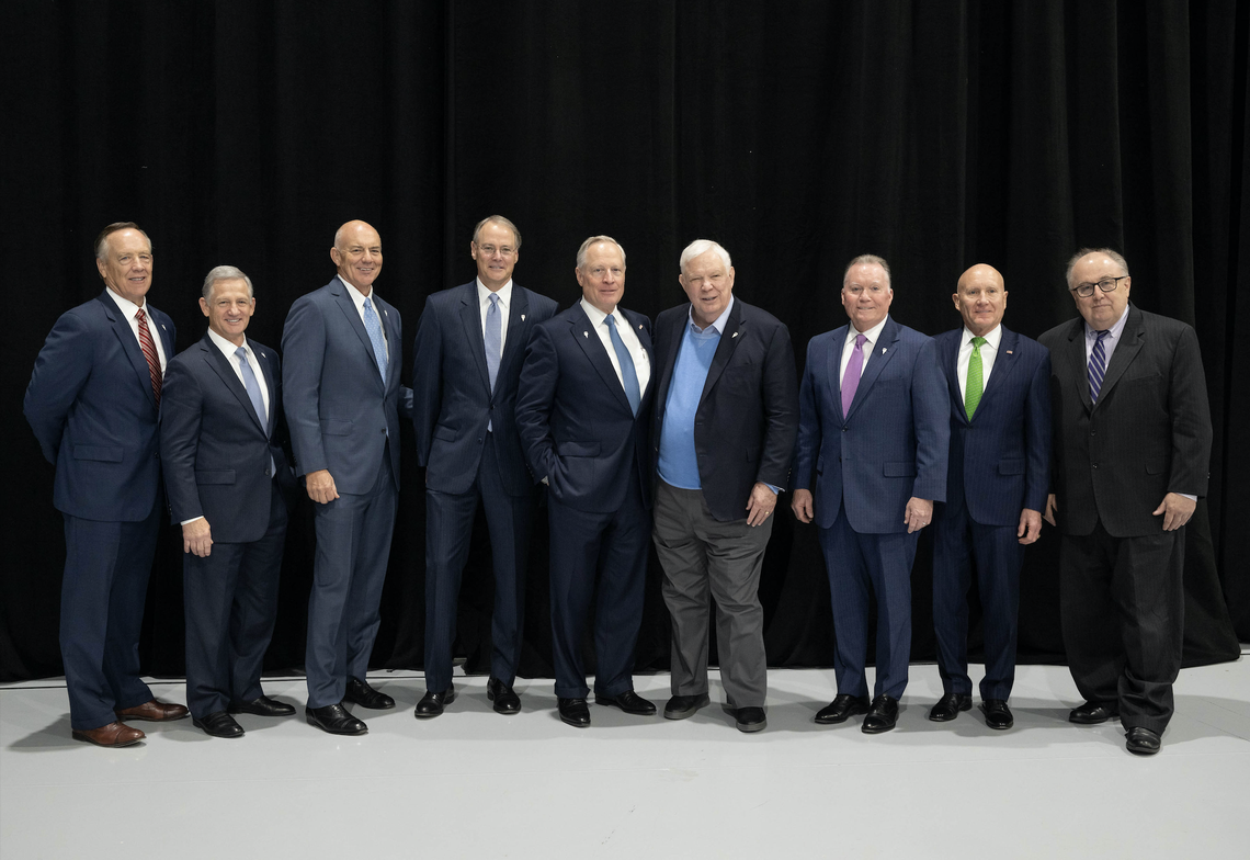 Nine men dressed in business suits stand and pose for a picture against a black curtain backdrop.