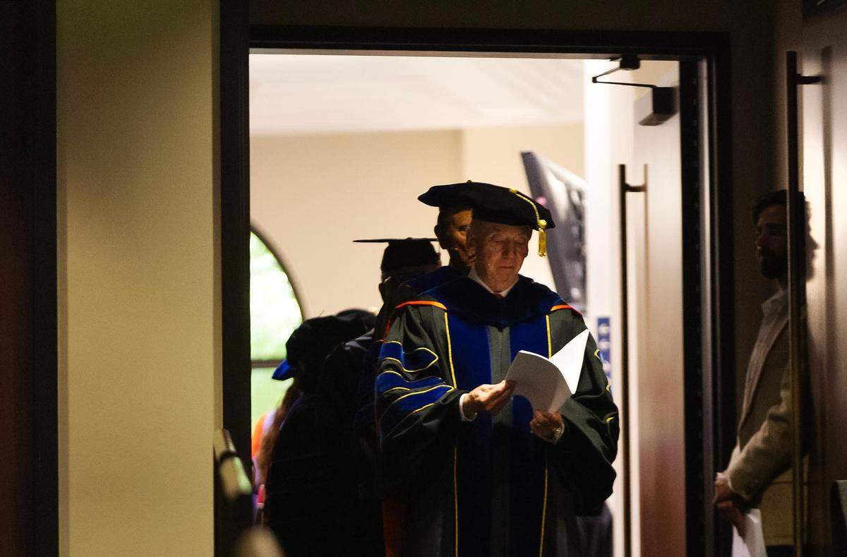A man in professor cap and gown looks down at notes while framed in a doorway.