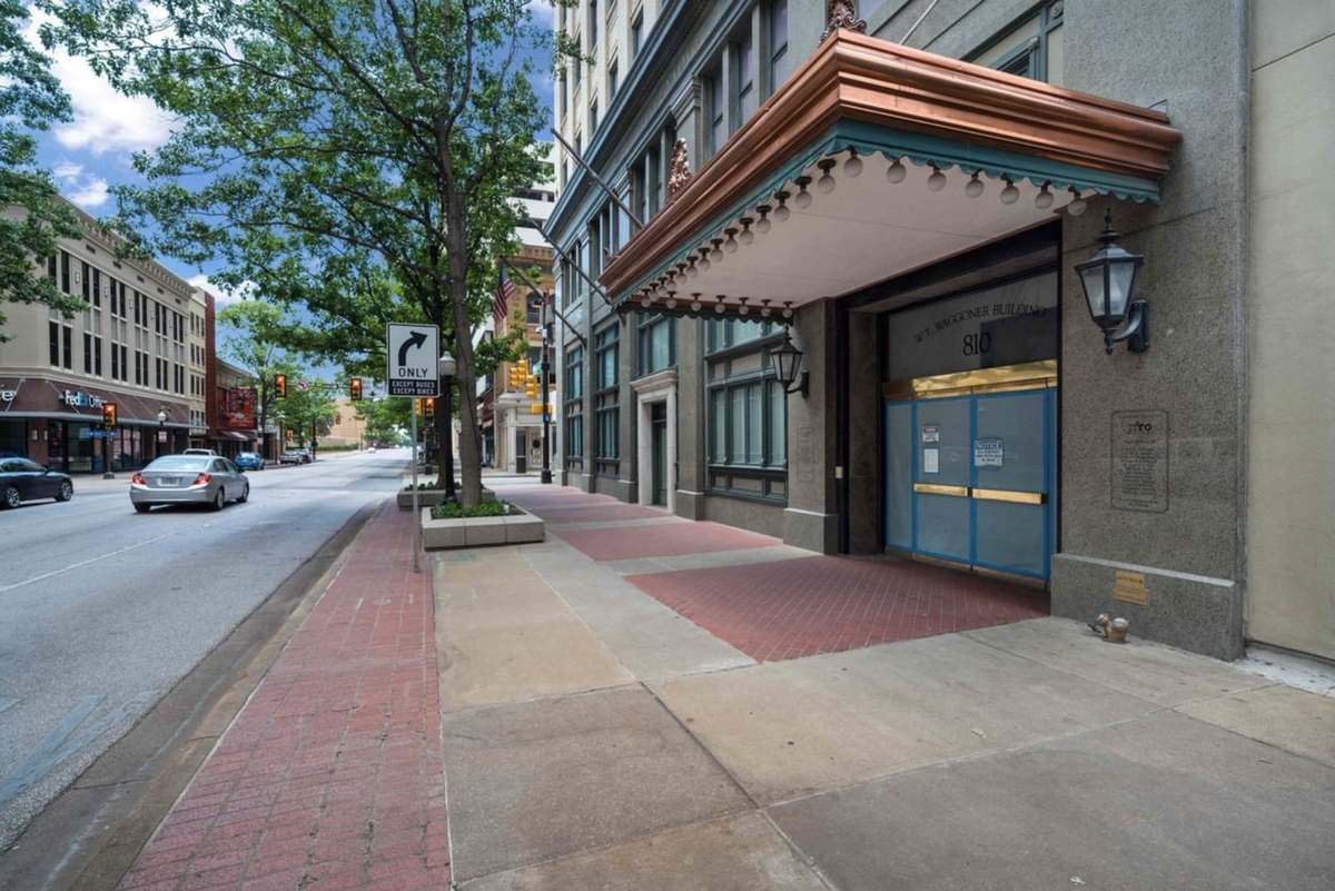 Red brick and gray concrete line the street outside the historic "Waggoner Building" previously transformed into the Sandman Signature Hotel. The hotel front features a bronze and turquoise awning with lights.