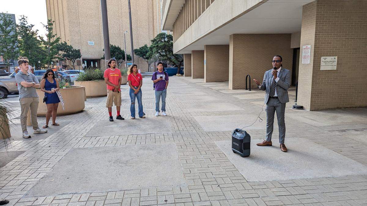 A man speaking in front of a concrete building to about 15 people