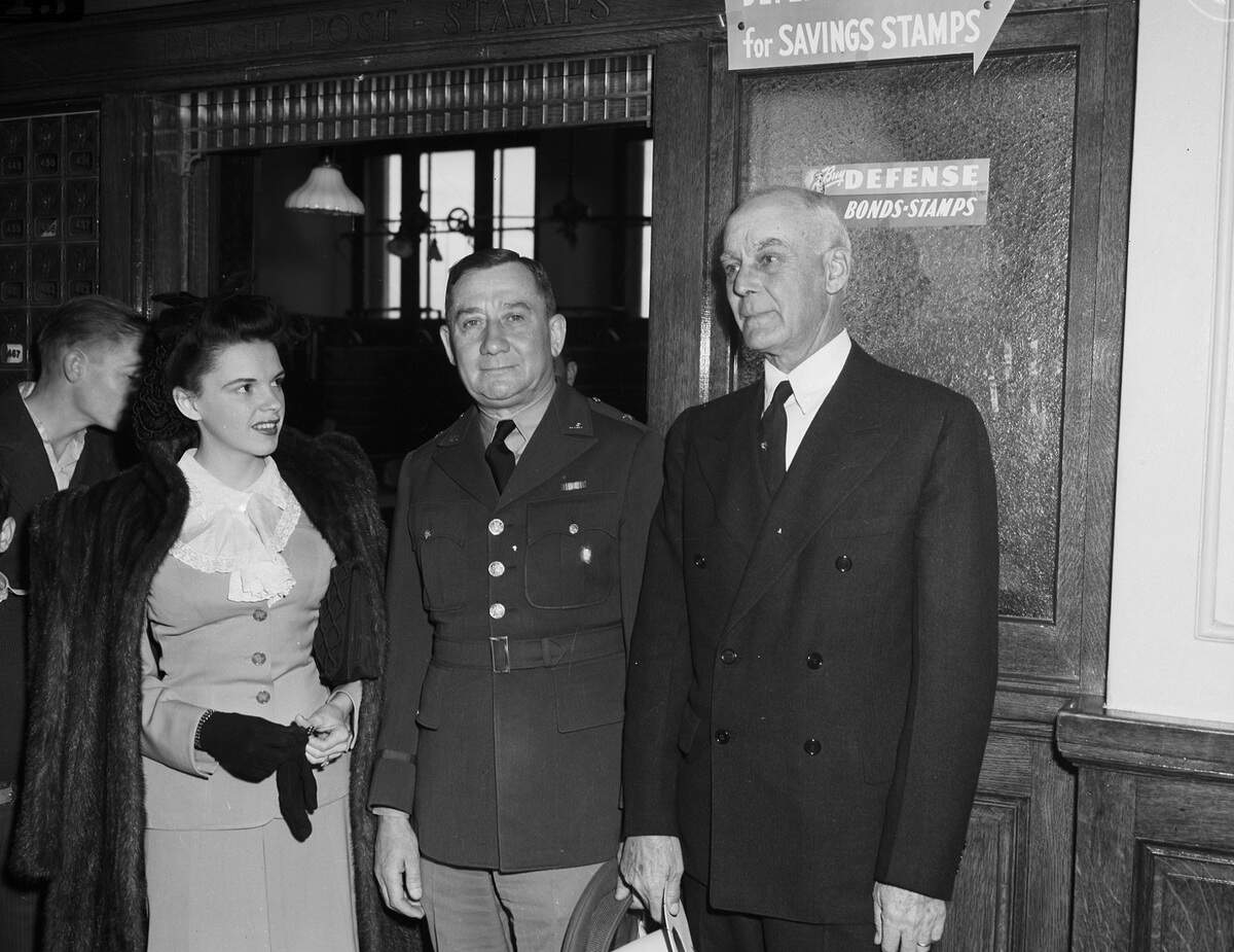 Garland is wearing a bow in her hair, a suit with lace around the collar, and a fur jacket over her shoulders. General Reinhart is the commander of Camp Wolters. He is dressed in his military uniform. Mr. Miller is the Mayor of Mineral Wells, Texas. He is dressed in a double breasted suit and holding his hat. The men were the reception committee and received the children purchasing defense stamps are the post office.